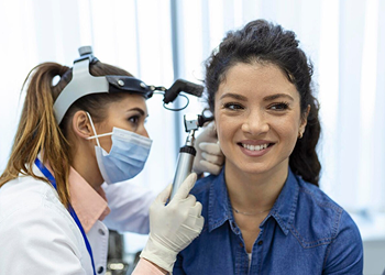Female doctor wearing a mask and headlamp examines a smiling woman’s ear using an otoscope during a medical check-up.