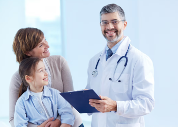 A smiling doctor with a clipboard stands with a happy mother and daughter in a bright clinic